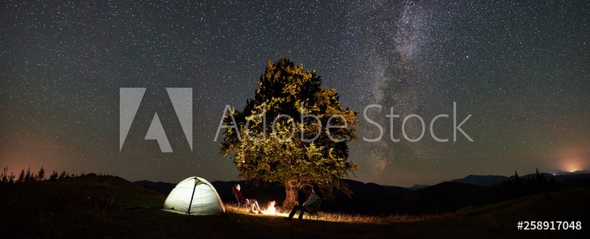 Picture of Panoramic view of couple tourists resting at summer night camping in mountains Travelers sitting on chairs beside campfire illuminated tent and big tree under starry sky full of stars and Milky way